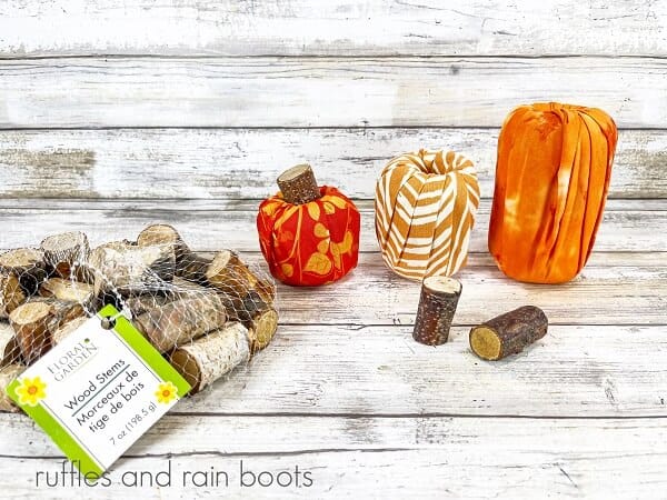 Three wrapped pool noodle pumpkins, one with a wooden stem, next to a bag of wooden stems against a white weathered wood background.