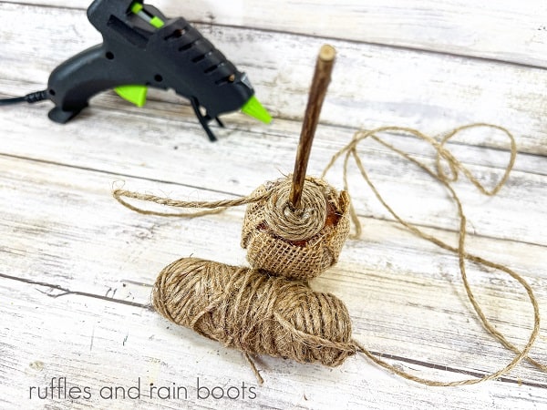 A Dollar Tree acorn being wrapped with jute twine, next to a ball of twine and a black hot glue gun against a white weathered wood background.