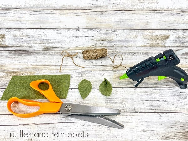 Green felt scraps, some cut into leaves next to a hot glue gun, twine and pinking shears against a white weathered wood background.