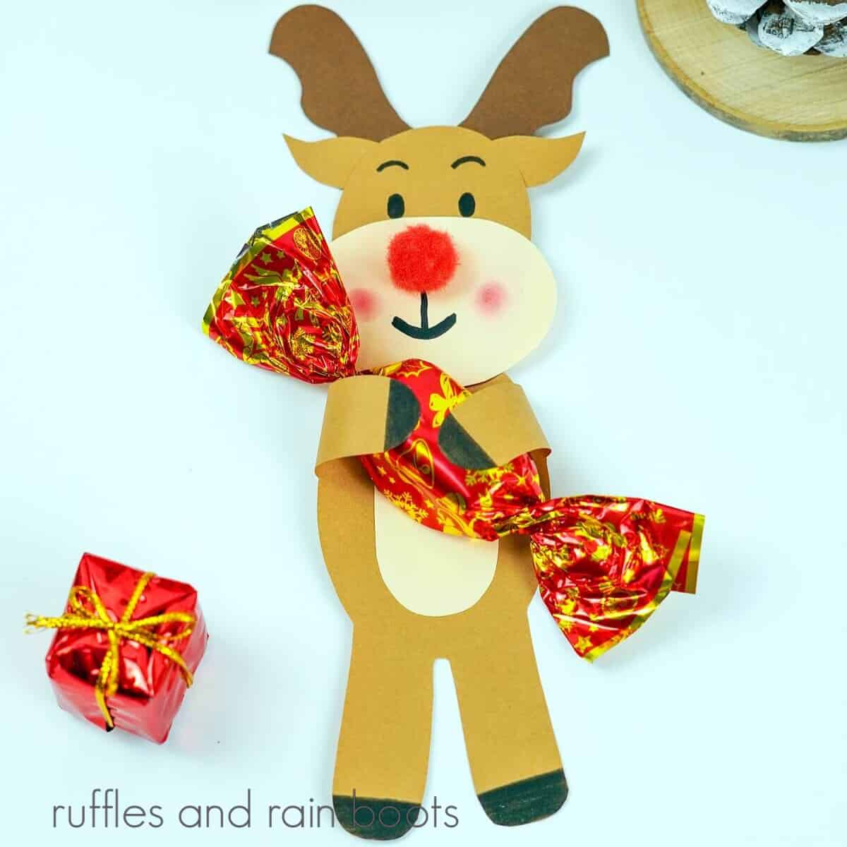 A square image of a paper cut-out reindeer holding a piece of candy next to holiday Christmas decorations against a white background.