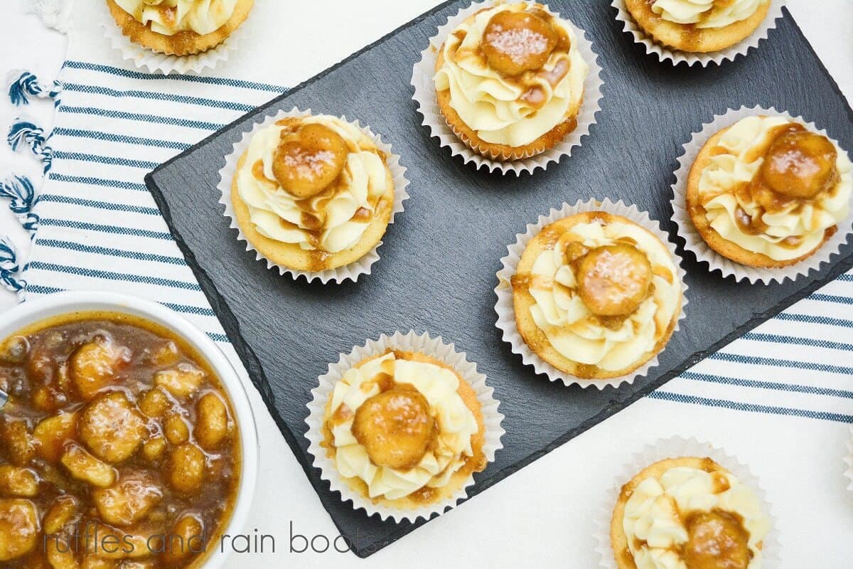 Six Bananas Foster Cupcakes on a piece of grey slate, next to a bowl of Bananas Foster on a white and blue stripe towel.