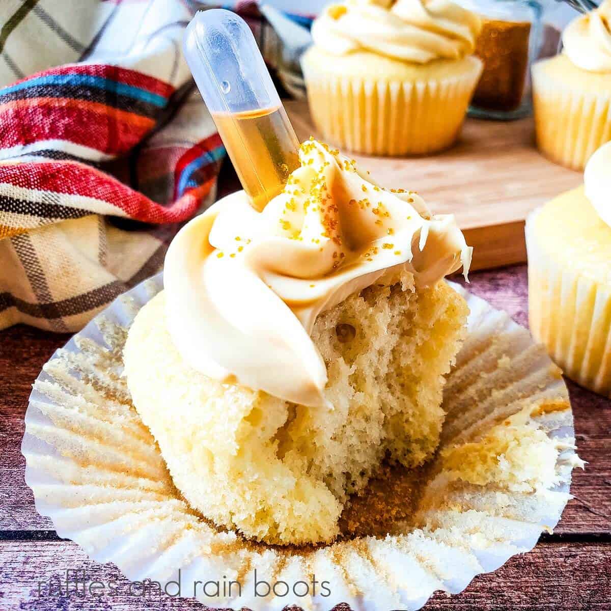 A cognac cupcake cut in half in front of a wooden cutting board with additional cupcakes on it, next to a plaid towel against a weathered wood background.