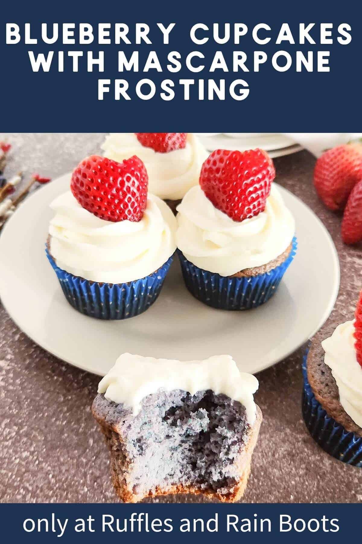 A plate of blueberry cupcakes with mascarpone frosting and fresh strawberries on top next to a blueberry cupcake with a bite taken out against a brown background.