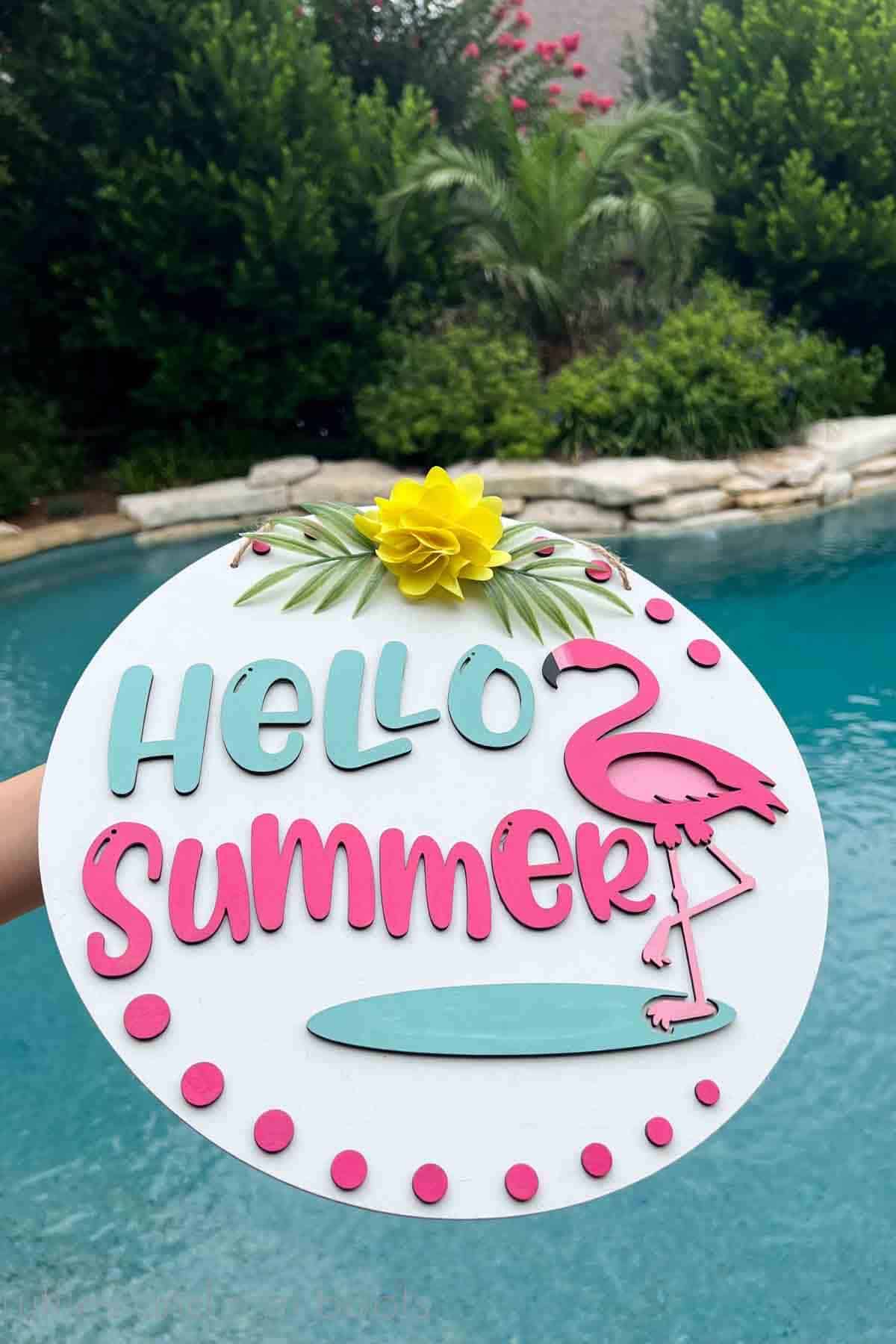 Vertical image of a white door sign for summer with flamingo accent held up in front of a tropical background and pool.