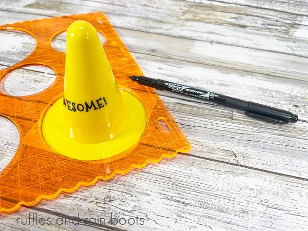 A plastic cone under a circle template next to a black magic marker against a white weathered wood background.