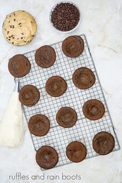 A metal baking rack with unfrosted cupcakes with small holes in the top, next to a bowl of edible cookie dough and a bowl of mini chocolate chips.