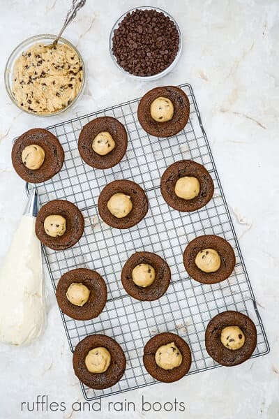 A metal baking rack with unfrosted cupcakes with edible cookie dough on top, next to a bowl of edible cookie dough and a bowl of mini chocolate chips.