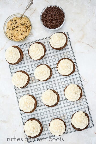 A metal baking rack with frosted cupcakes, next to a bowl of edible cookie dough and a bowl of mini chocolate chips.