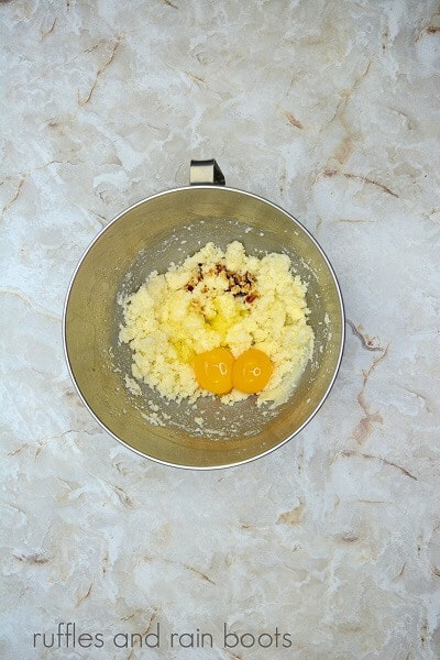 A large metal mixing bowl filled cupcake batter and an egg and an egg yolk, against a marble surface.