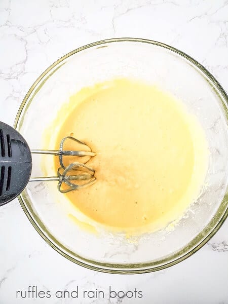 Beaten cupcake batter in a round glass bowl, with electric metal beaters against a white marble surface.