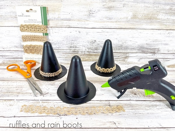 Three cones painted black, 2 with burlap trim, next to a package of burlap tri, a pair of scissors and a hot glue gun against a white weathered wood background.