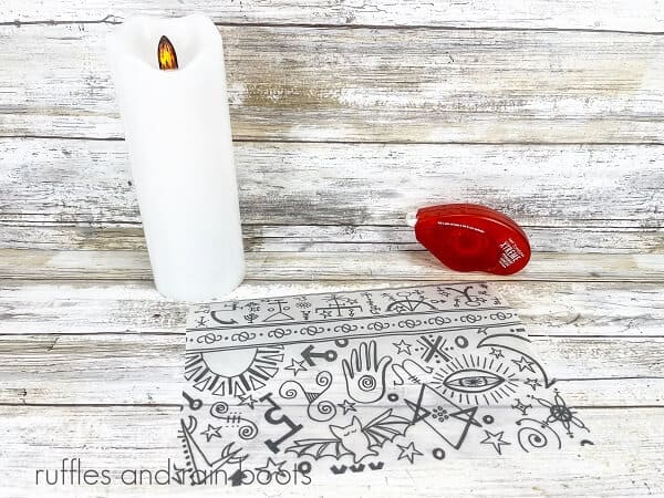 A plain battery operated candle, next to the free pattern and a roll of adhesive against a white weathered background.