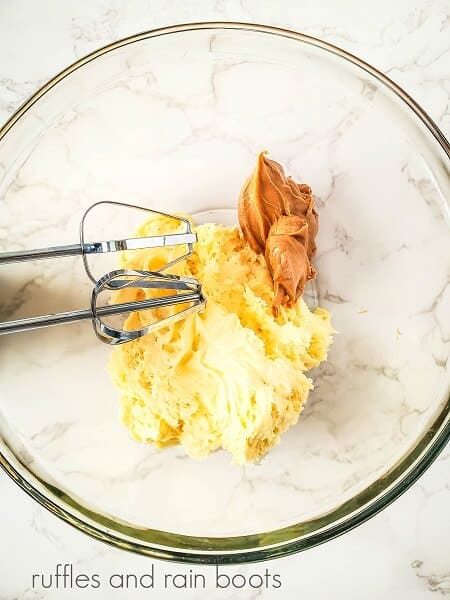 Frosting and peanut butter in a large round glass bowl with electric beaters on the inside of the bowl on a white marble surface.