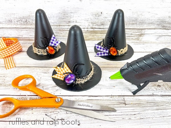 Three completed witch's hats, next to orange gingham ribbon, a pair of scissors and a hot glue A completely decorated black witch's hat in the foreground, next to a witch's hat on a yellow gourd, next to a second gourd against a white weathered wood background.