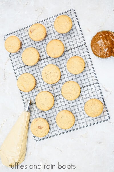 Baked cupcakes on a wire cooling rack with dulce de leche and a piping bag full of frosting on the side on top of a white marble surface.