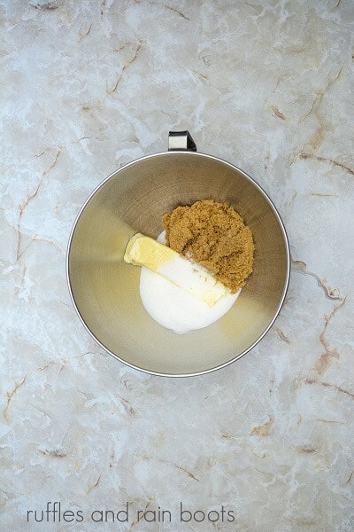 A large metal mixing bowl filled with butter, sugar, brown sugar and vanilla, against a marble surface.