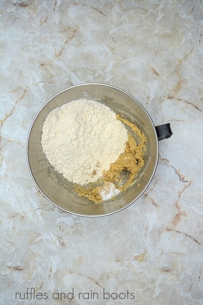 A large metal mixing bowl filled with edible cookie dough and flour against a marble surface.