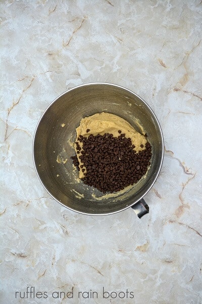 A large metal mixing bowl filled with edible cookie dough and mini chocolate chips against a marble surface.