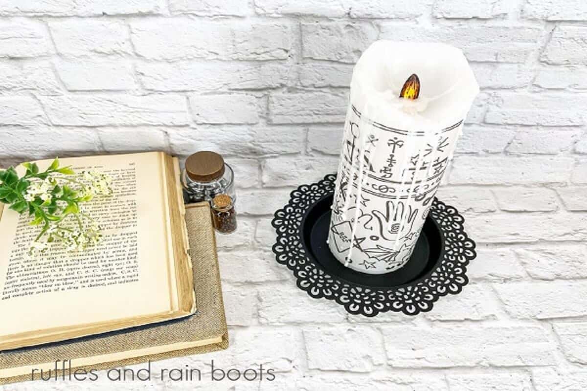 A horizontal image of a Hocus Pocus Black Flame Candle on a black candle holder, next to a few potion bottles and two books, one open with faux greenery on top on the left against a white brick background.