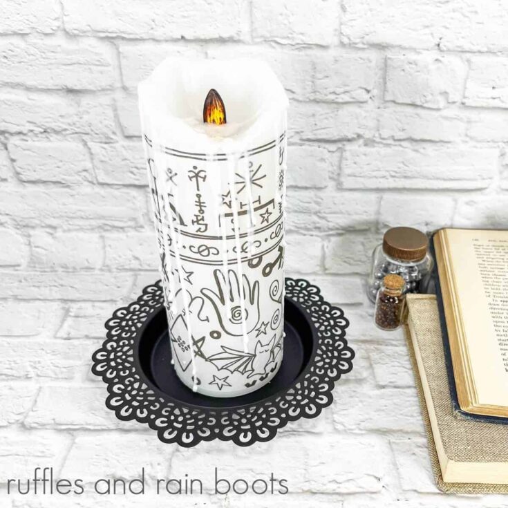 A vertical image of a Hocus Pocus Black Flame Candle on a black candle holder, next to a few potion bottles and two books, one open with faux greenery on top on the right against a white brick background.