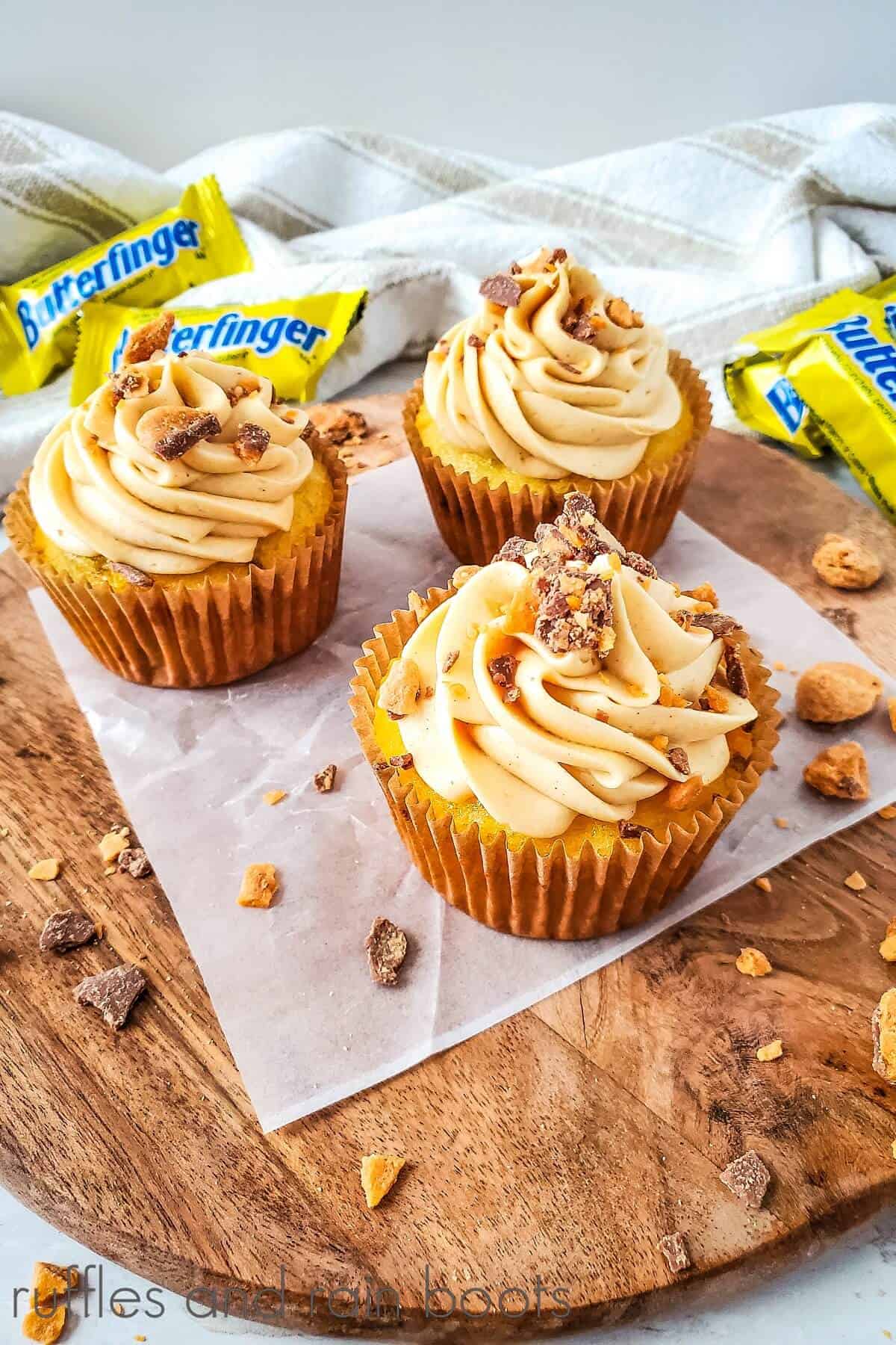 3 Butterfinger cupcakes on a wooden cutting board on a piece of parchment paper with a white towel and Butterfinger candy bars in the background.