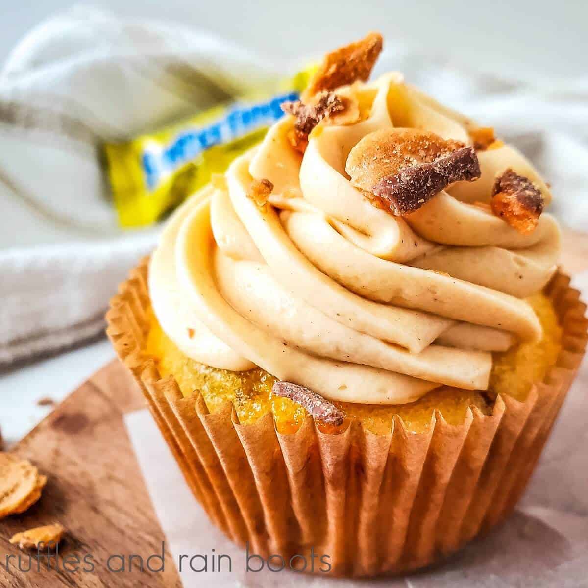 A square close up of a Butterfinger cupcake on a wooden cutting board with a Butterfinger candy bar in the background next to a white towel.