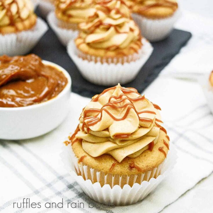 A square image of a close up of a dulce de leche cupcake, net to a bowl of dulce de leche nearby and a tray of cupcakes on a white and blue plaid towel.