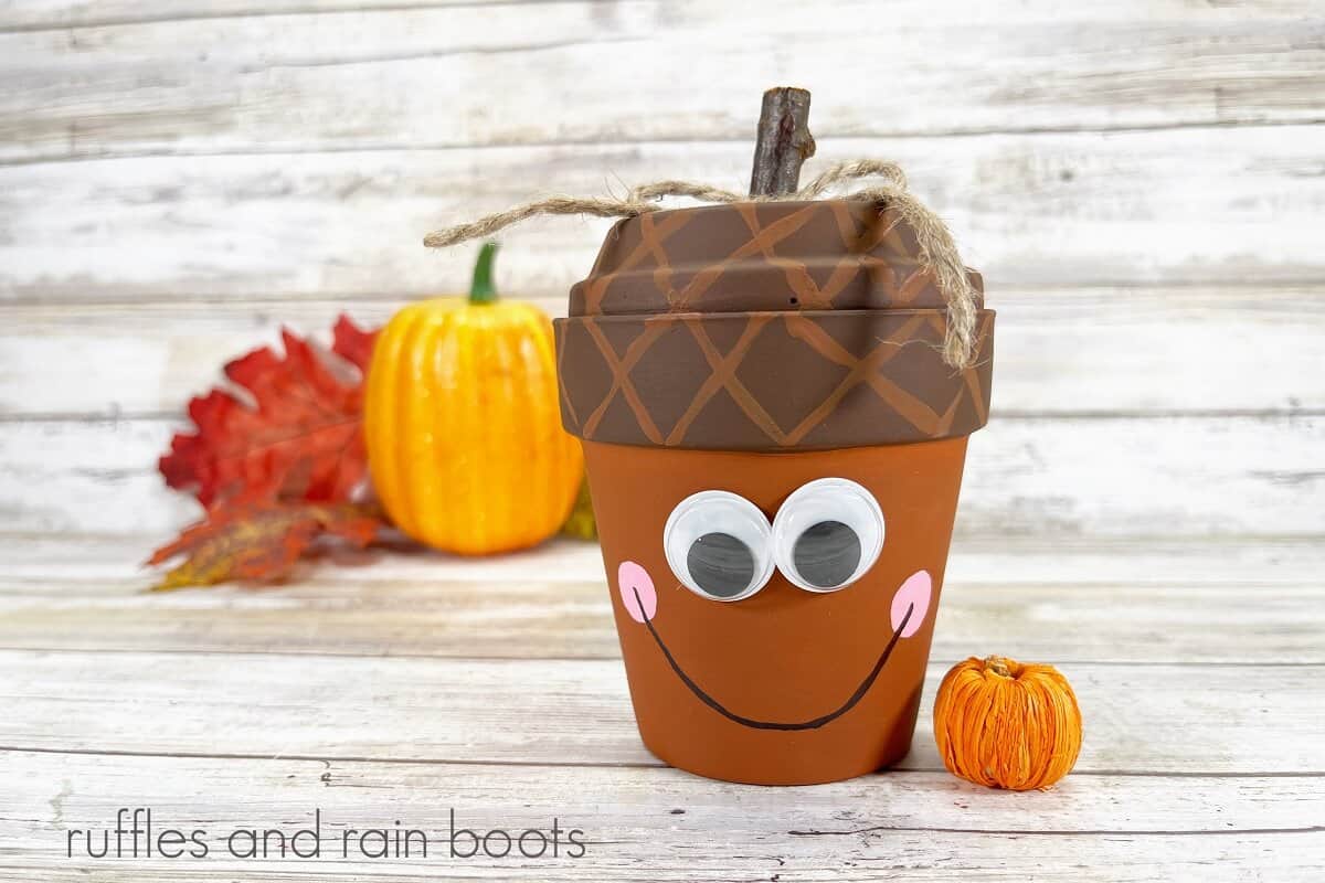A horizontal close up of a clay pot acorn craft for fall next to two faux pumpkins and fall leaves on a weathered white wood background.