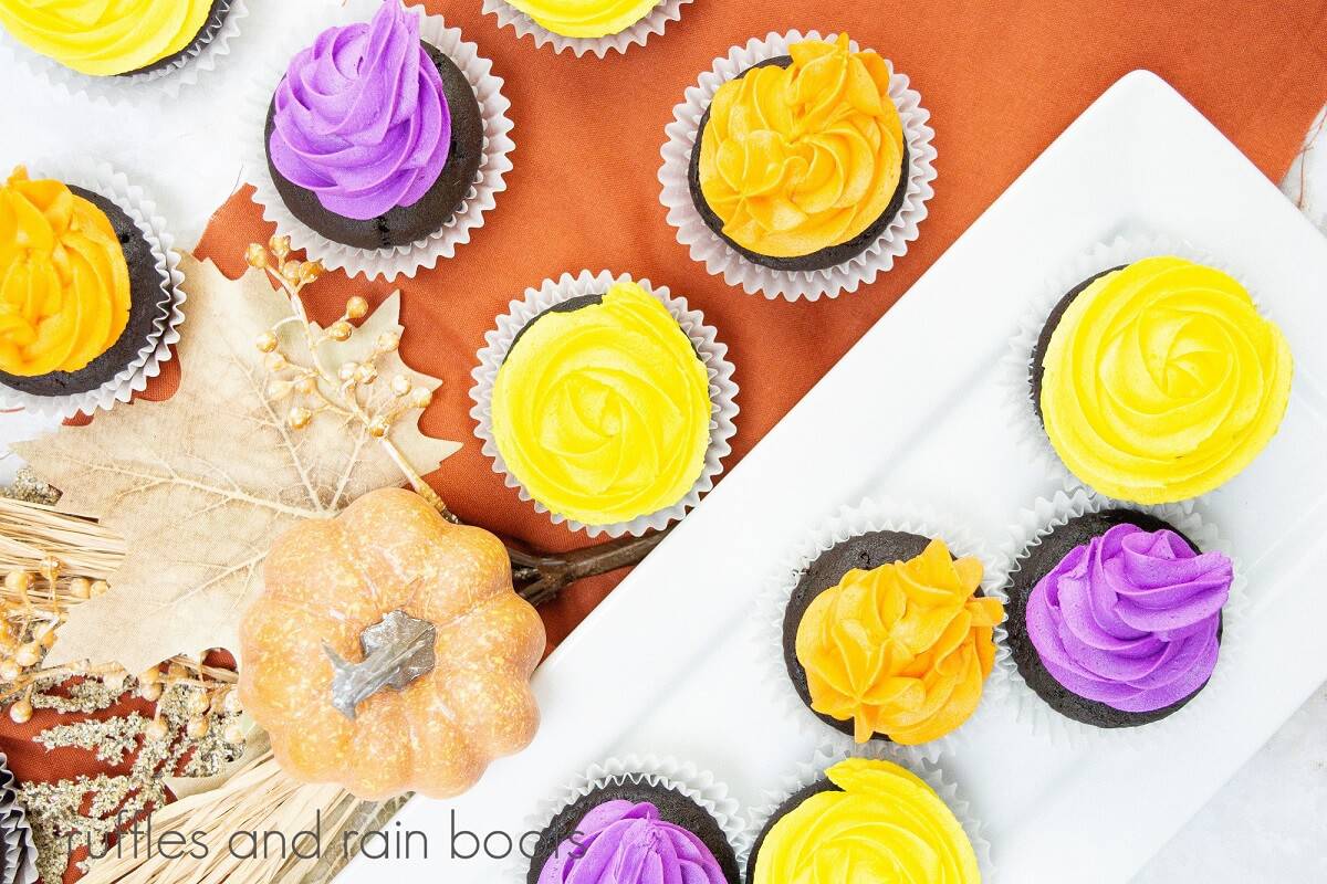 An overhead image of a rectangular white plate full of Hocus Pocus cupcakes, with more cupcakes on a burnt orange napkin, next to faux fall decorations.