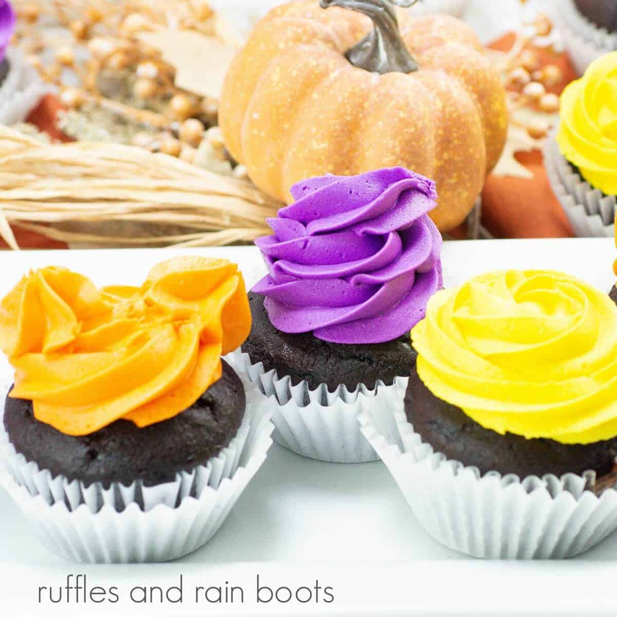 Three frosted cupcakes for the Hocus Pocus colors of yellow, orange and purple, on a white plate and next to a faux pumpkin and other fall decorations against a white marble background.