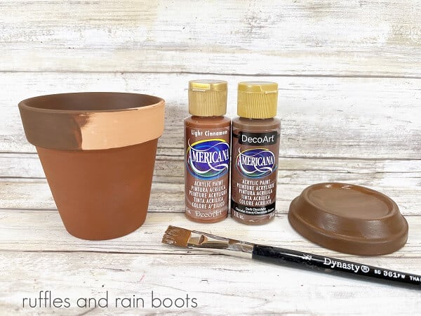 A painted clay pot next to two bottles of brown paint, a painted saucer and a paintbrush on a weathered white wood background.