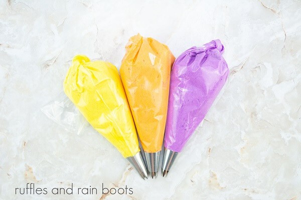 Piping bags filled with yellow, orange and purple frosting against a white marble background.