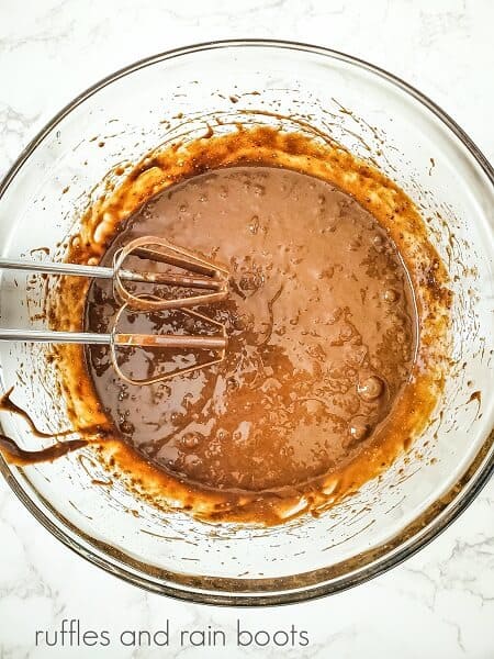 A large mixing bowl with mixed chocolate cake batter with electric beaters inside on a white marble surface.