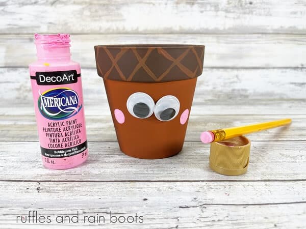 A clay pot acorn craft with googly eyes, next to an open bottle of pink craft paint and a pencil, with the eraser dipped into the paint on a weathered white wood background.