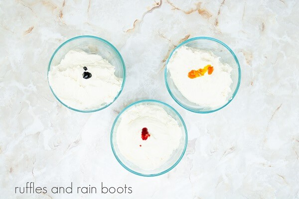 Three bowls of white frosting with drops of food coloring against a white marble background.