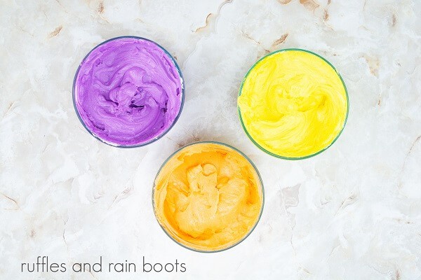 Three bowls filled with purple, yellow and orange frosting against a white marble background.