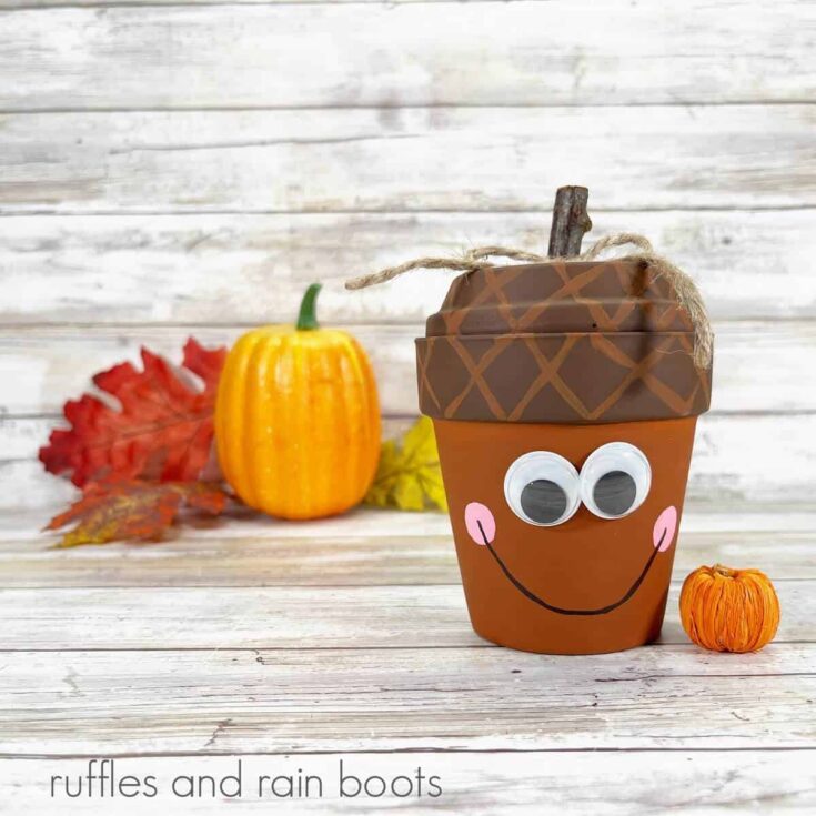 A square photo of a clay pot acorn craft for fall next to two faux pumpkins and fall leaves on a weathered white wood background.