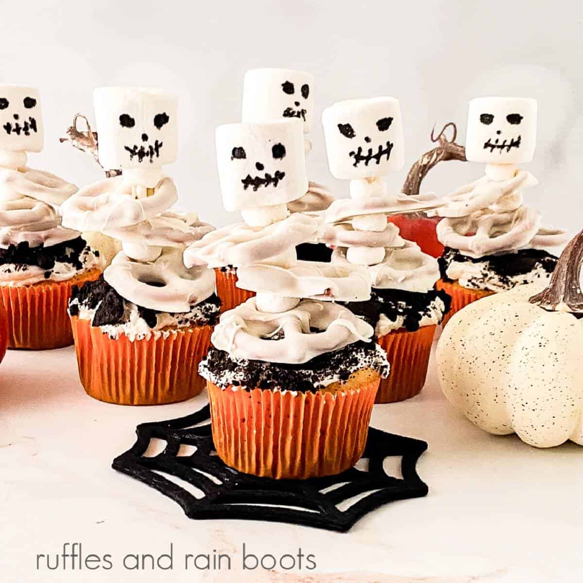 6 skeleton cupcakes next to a white faux pumpkin against a white background.