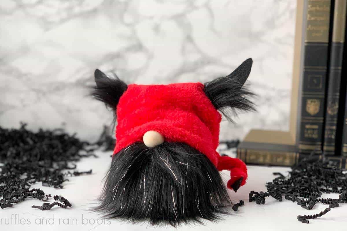 Horizontal image of a red devil gnome with a black gnome beard with tinsel placed in front of marble background with old books.