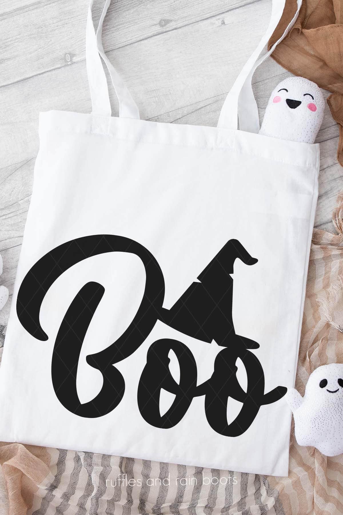 Vertical image of a white tote bag on neutral background with cute ghost plushies which reads boo in black vinyl with a witch hat.