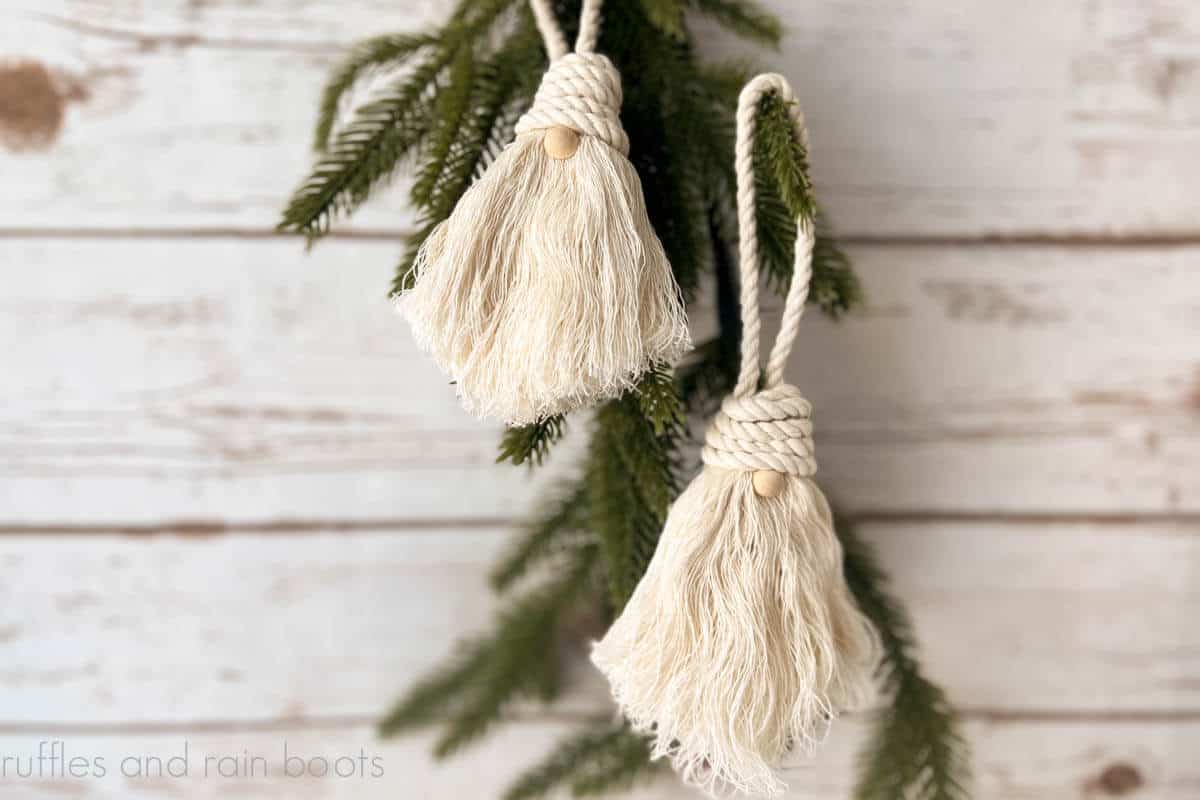 Horizontal image of two natural colored rope gnomes hanging on pine branches in front of a white wood wall background.