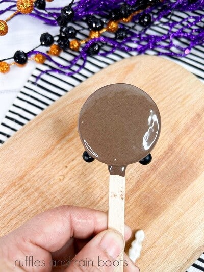 An Oreo dipped in melted chocolate against a wooden cutting board on a black and white striped towel with Halloween decorations in the background