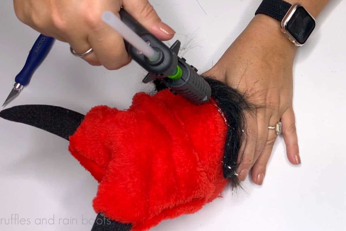 Crafter using a hot glue gun to secure a faux fur gnome beard to a devil gnome body.