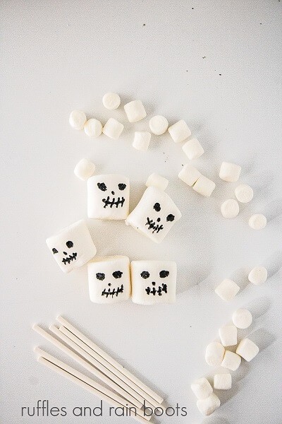 Several mini marshmallows next to 5 standard marshmallows with faces drawn on them and 6 lollipop sticks against a white background.