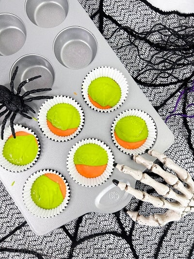 A green and orange Halloween colored cupcake batter in a cupcake pan with a fake skeleton hand and spider on the pan on a sheer spiderweb covering