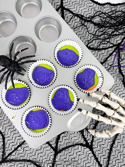 A purple, green and orange Halloween colored cupcake batter in a cupcake pan with a fake skeleton hand and spider on the pan on a sheer spiderweb covering