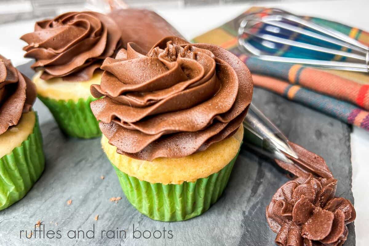 Three frosted cupcakes with chocolate frosting next to a piping bag filled with chocolate frosting on a grey marble slab next to a whisk and a plaid towel against a kitchen backsplash.