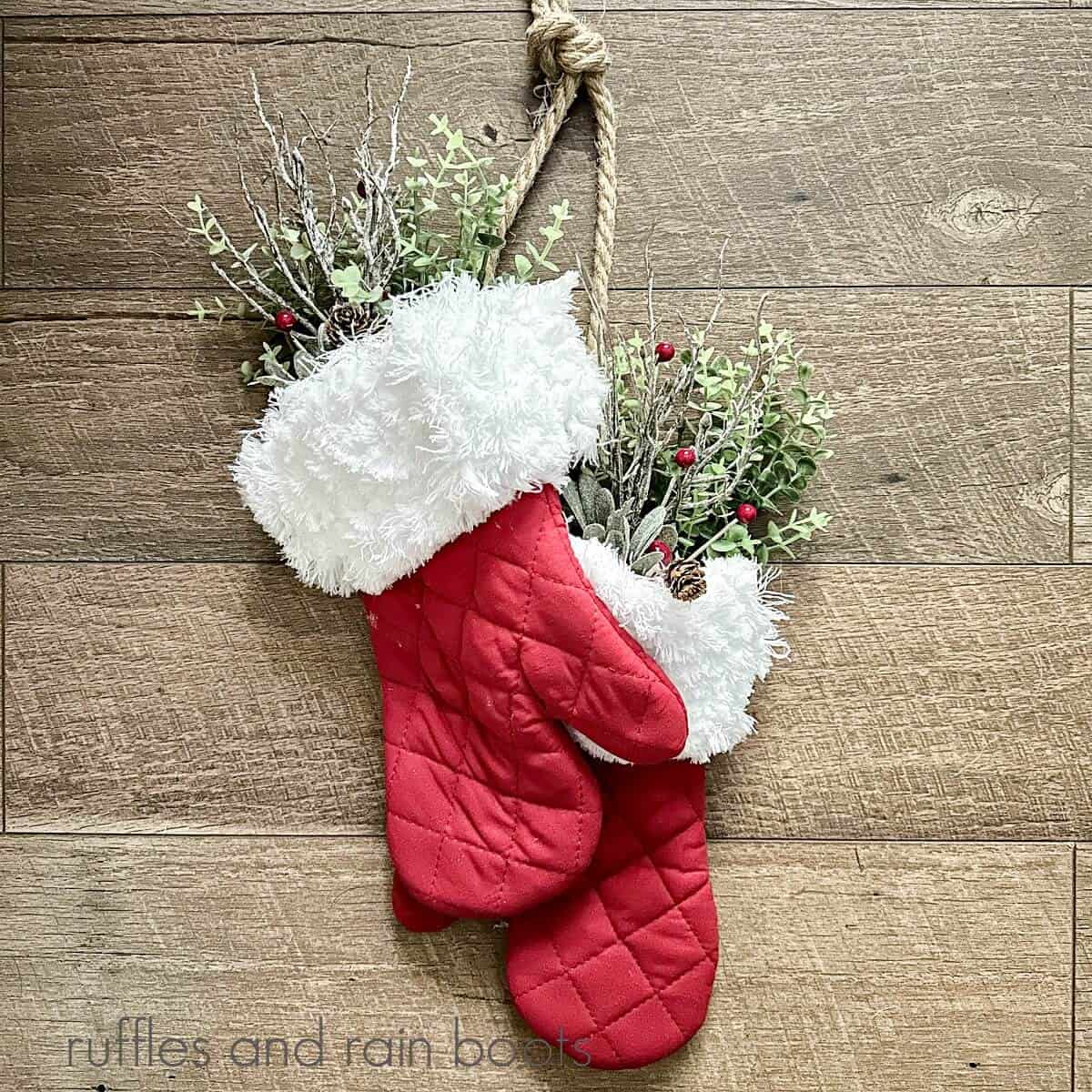 A close up image of a pair of red oven mitts made into Santa mittens with greenery inside hanging against a wooden surface.