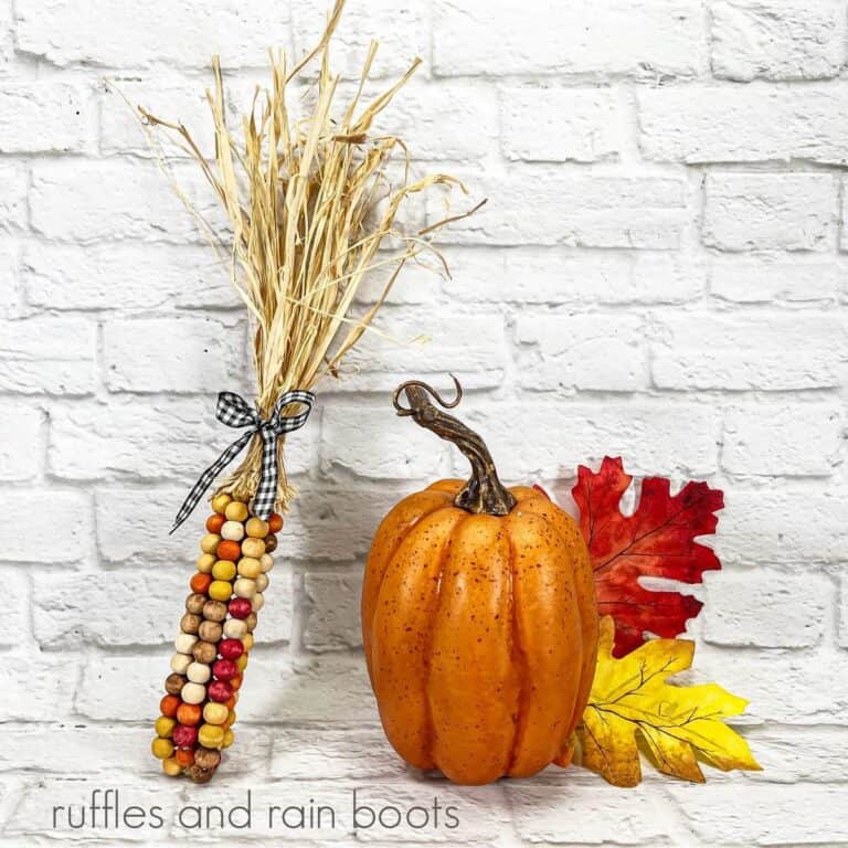 A square image of a bunch of fall harvest corn standing vertically with a black and white gingham ribbon, next to a faux pumpkin and fall colored leaves against a white brick backdrop.