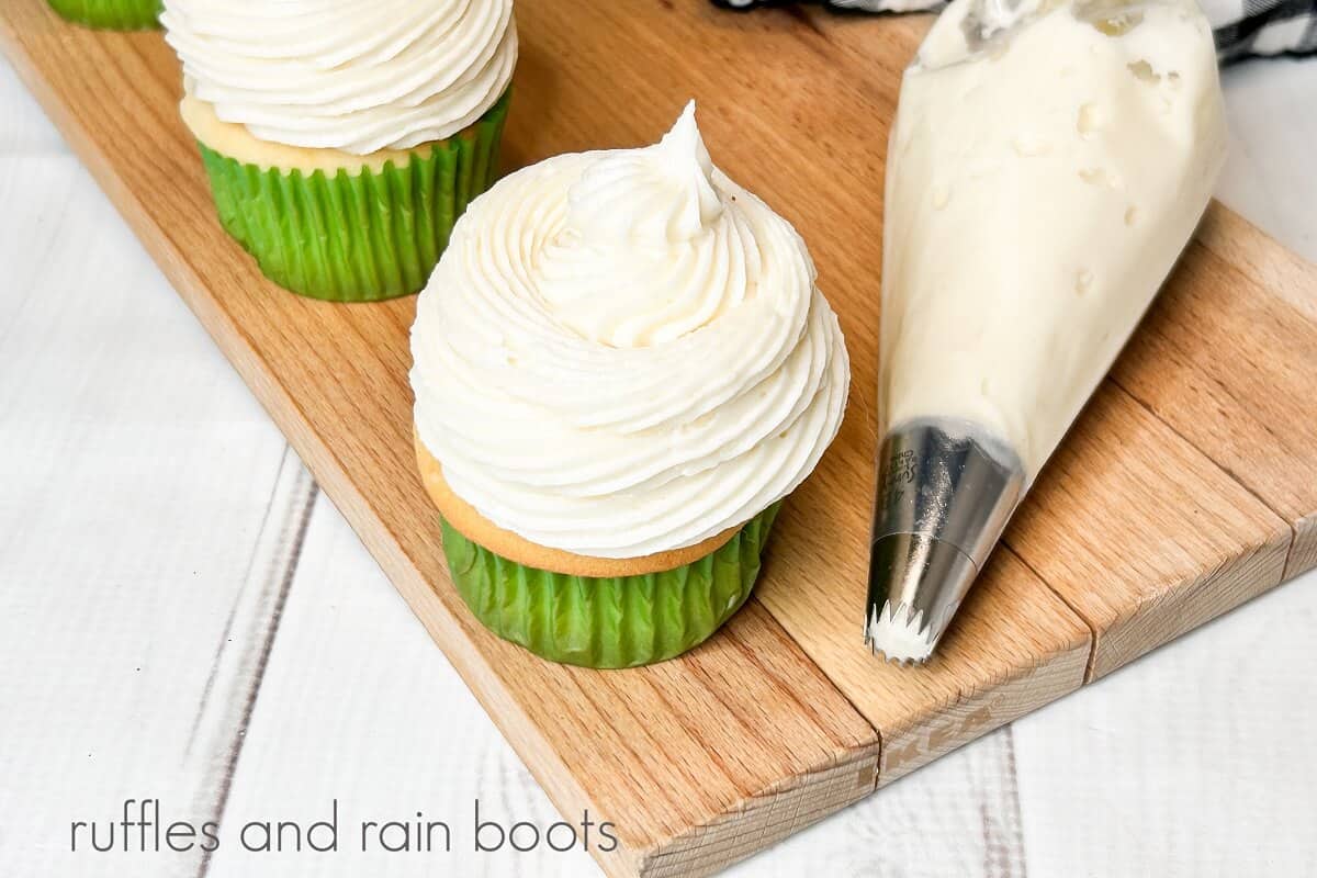 Two frosted vanilla cupcakes with a piping bag filled with easy cream cheese frosting on a wooden cutting board with a black and white checked towel in the background on a white wooden backdrop.